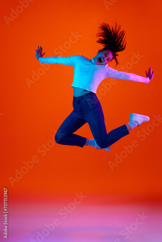 Full length portrait of young overjoyed woman jumping in motion of joy and happiness against gradient pink-orange background in neon light. Concept of self-expression, beauty and fashion, energy. Ad