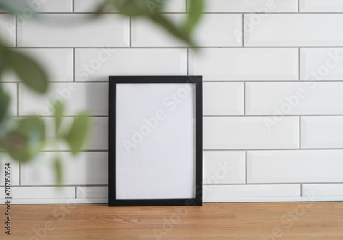 A black frame with a blank canvas against a white tile wall and on a wooden tabletop with blurred green foliage.