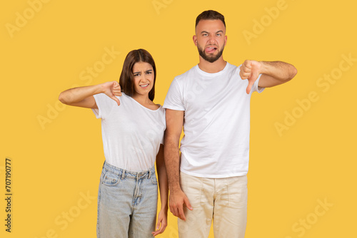 Disapproving young couple in white t-shirts giving thumbs down gestures with displeased expressions