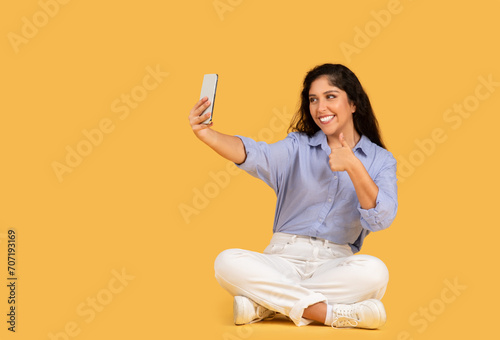 A cheerful young woman in casual clothes sits cross-legged on a yellow background