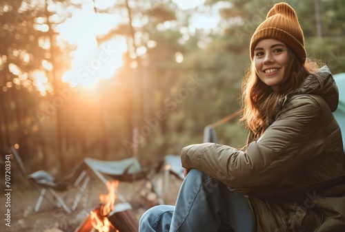 Beautiful young woman with brunette hair camping in the forest wilderness, sitting in a camping chair, smiling and looking at the camera. Enjoying morning sunshine in the woods, happy female camper photo