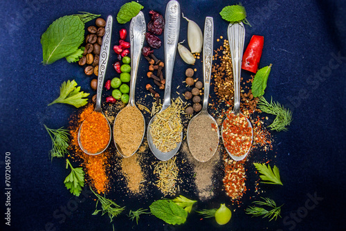 Fototapeta Naklejka Na Ścianę i Meble -  Five spoons with spices lie next to coffee, chili peppers and pomegranate seeds on a dark background next to peas, parsley, dill and green leaves in close-up	
