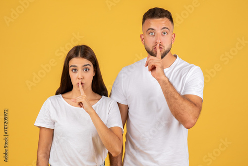 Surprised young couple in white t-shirts with fingers on lips making a shush gesture