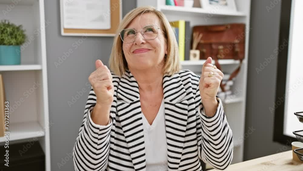 Radiant middle-aged blonde business woman ecstatically celebrates a victorious win, her smile screams triumph, her expression oozes joy in the office setting. Radiant middle-aged blonde business woman ecstatically celebrates a victorious win, her smile screams triumph, her expression oozes joy in the office setting.