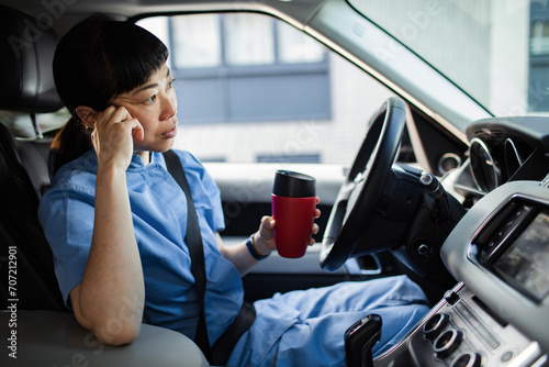 Woman in scrubs looking tired holding coffee mug in car