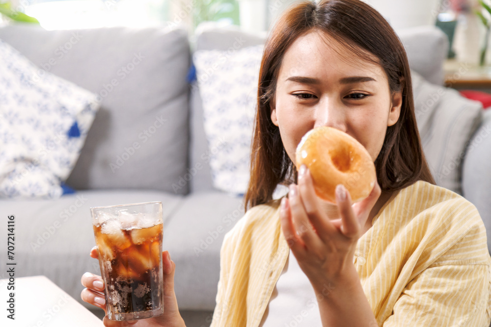 young asian woman enjoy eating sweet donut and drink cola soft drink in living room at home in summer season.
