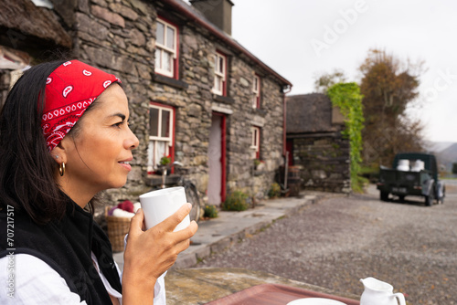 Beautiful latin woman holding cup of coffee while resting next to beautiful stone cottage in the Irish hinterland on her rural road trip photo
