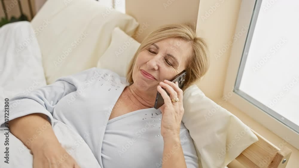 A smiling middle-aged woman talks on the phone while relaxing in her bedroom, showcasing a comfortable, casual lifestyle moment. A smiling middle-aged woman talks on the phone while relaxing in her bedroom, showcasing a comfortable, casual lifestyle moment.
