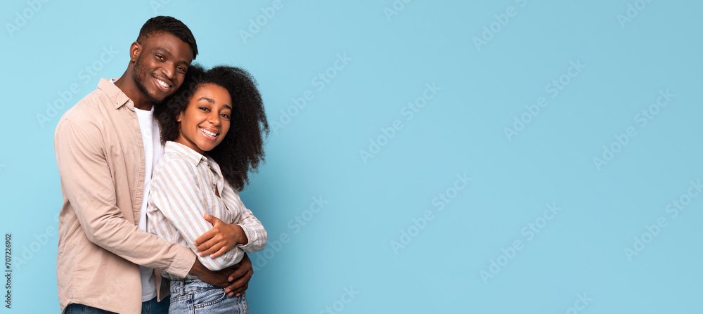 Fototapeta premium Affectionate black couple hugging and smiling on blue background, panorama