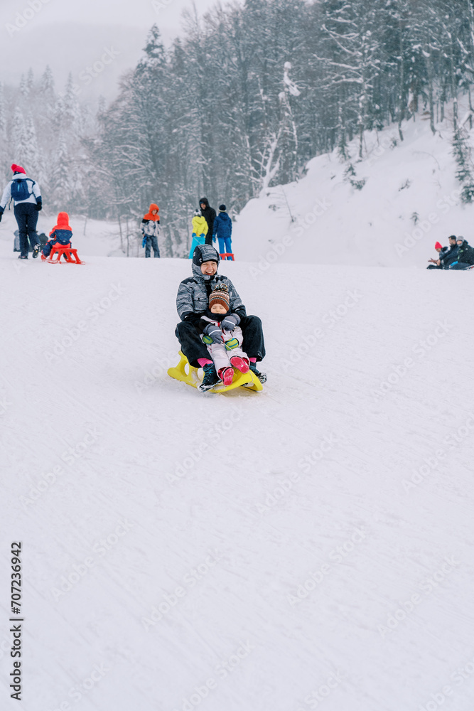 Smiling mother with a small child goes down the slope of a snowy mountain on a sled