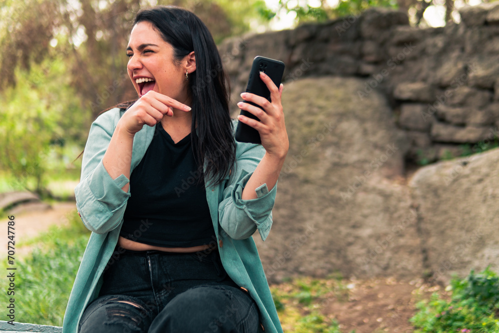 Beautiful Latin woman checking her phone and receiving good news, with an expression of joy, laughter, surprise, affection, etc. in an outdoor park