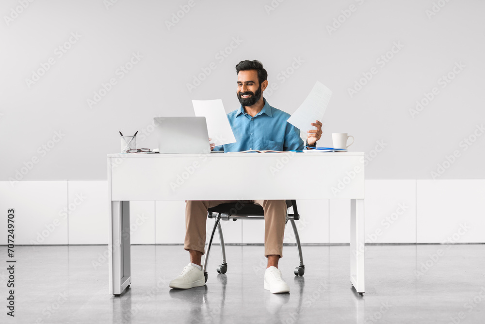 Happy indian professional man multitasking with documents and laptop Happy indian professional man multitasking with documents and laptop