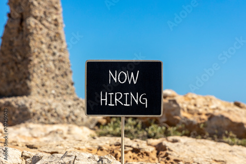 Now hiring symbol. Concept words Now hiring on beautiful black chalk blackboard. Beautiful brown stone blue sky background. Business marketing, motivational now hiring concept. Copy space.