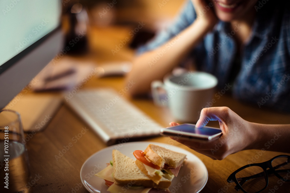 Woman smiling and using smartphone while having breakfast at home computer