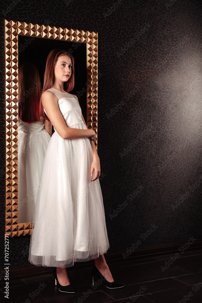 Full length of perfect cover teen girl in elegant white dress posing at big mirror, serious looking away. Chic teenage lady poses in shadow dark room. Youth gen Z emotion concept. Copy ad text space