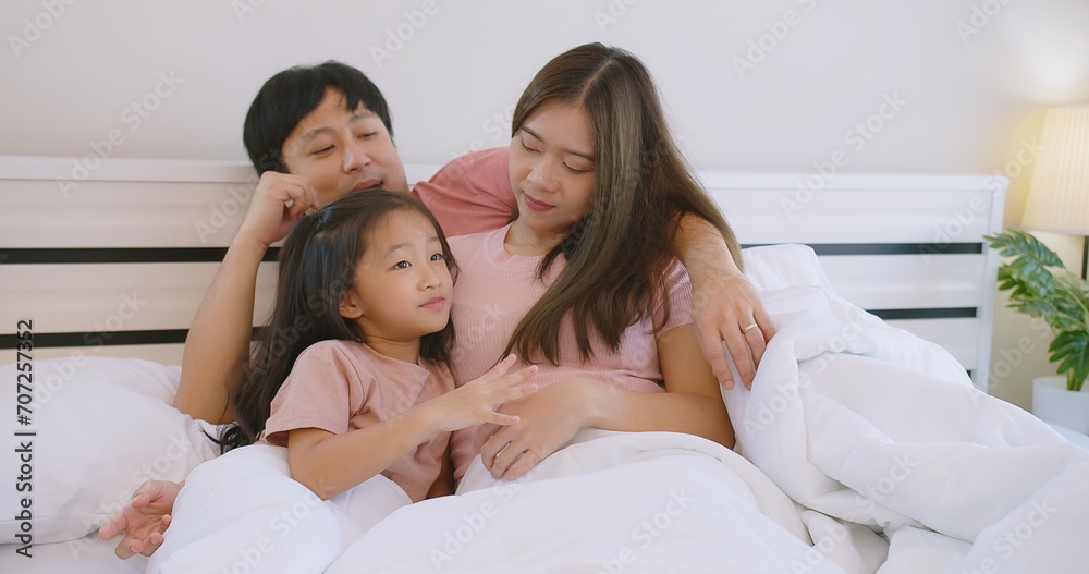 Happy Asian family father mother and daughter embrace smiling together lying relaxing on white bed in bedroom at home, happy parent and child girl enjoy hugging having fun playing in bedroom