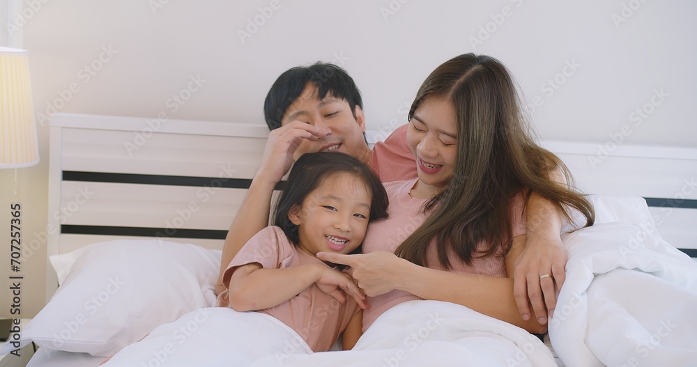 Happy Asian family father mother and daughter embrace smiling together lying relaxing on white bed in bedroom at home, happy parent and child girl enjoy hugging having fun playing in bedroom