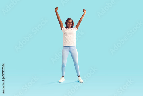 Young woman celebrates success after winning prize. Full body length happy joyful beautiful slim African American girl in casual wear standing on bright blue background, raising hands up and cheering