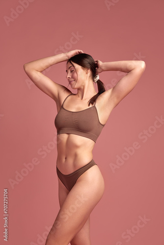 Happiness and wellness. Beautiful brunette young woman with slim healthy body standing in cozy cotton underwear over pink background. Concept of natural beauty, health and body care, body-positivity