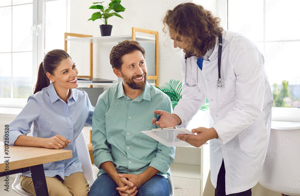 Obraz premium Happy young couple sitting at the desk and talking with a male family doctor therapist looking to report file with appointment during medical exam in clinic planning pregnancy. Health care concept.