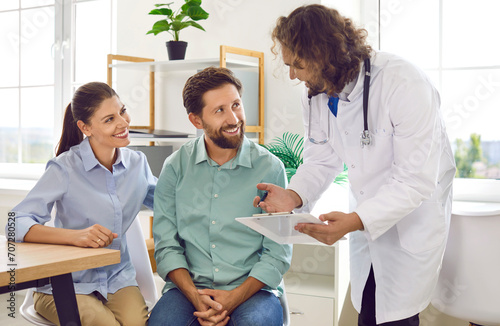 Happy young couple sitting at the desk and talking with a male family doctor therapist looking to report file with appointment during medical exam in clinic planning pregnancy. Health care concept.