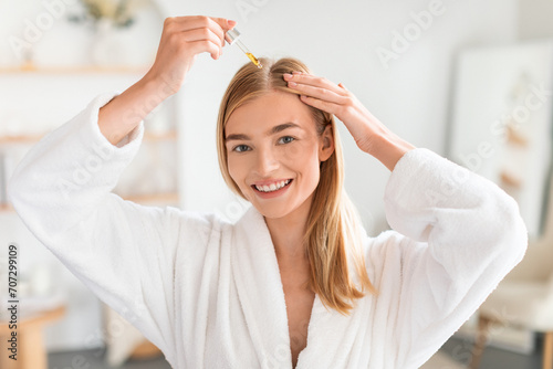 Joyful woman treats hair roots applying nourishing serum in bathroom photo