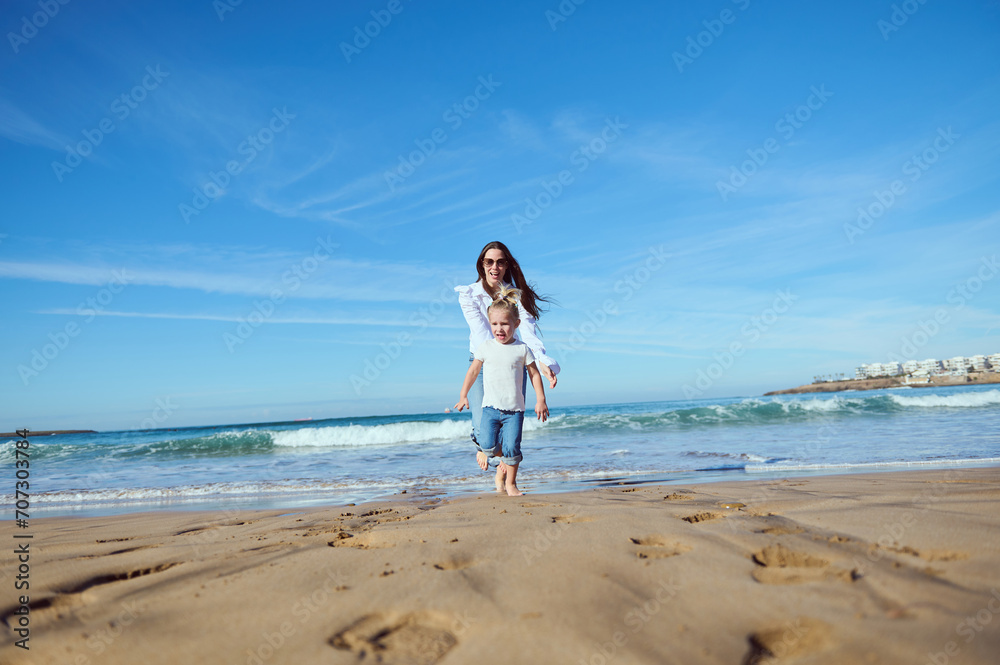 Cheerful happy mom catches her daughter while running barefoot, playing together on beach, leaving footsteps on wet sand