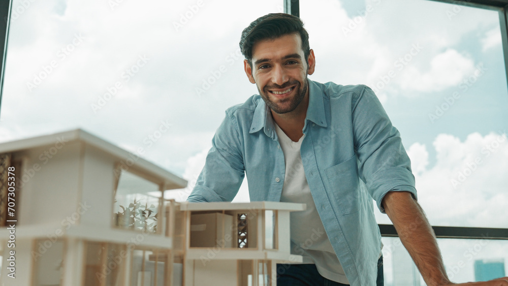 Portrait of engineer in casual outfit smiling at camera while inspect house model. Skilled architect looking at camera and standing near house model, project plan, architectural model. Tracery