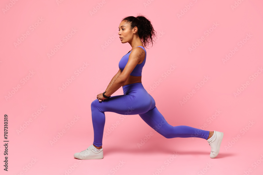 black woman in activewear performing forward lunges on pink backdrop