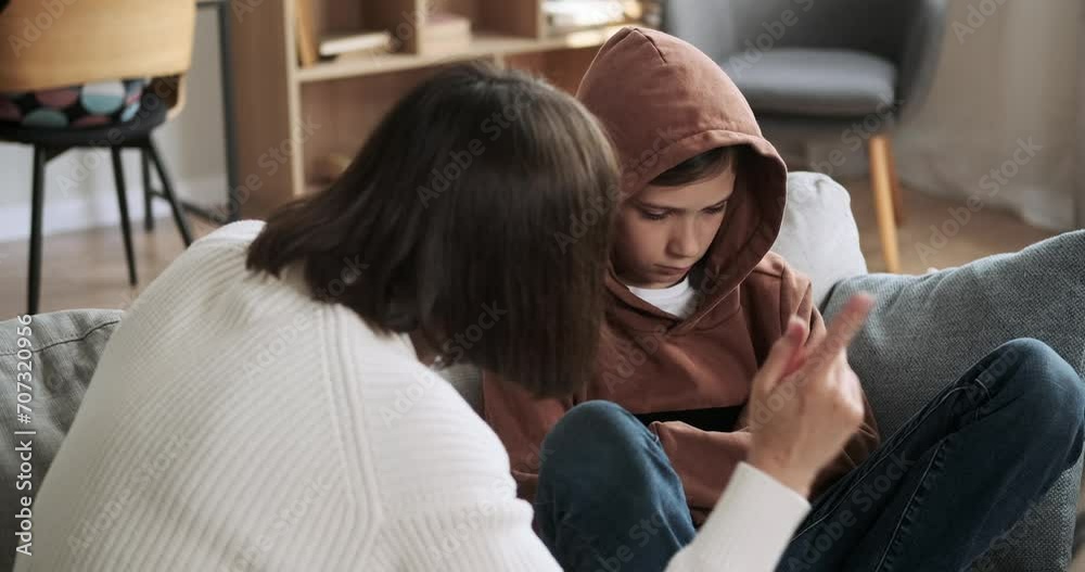 Unhappy schoolboy sits on the living room couch while receiving a scolding from his mother. The atmosphere is tense as they address an issue together.
