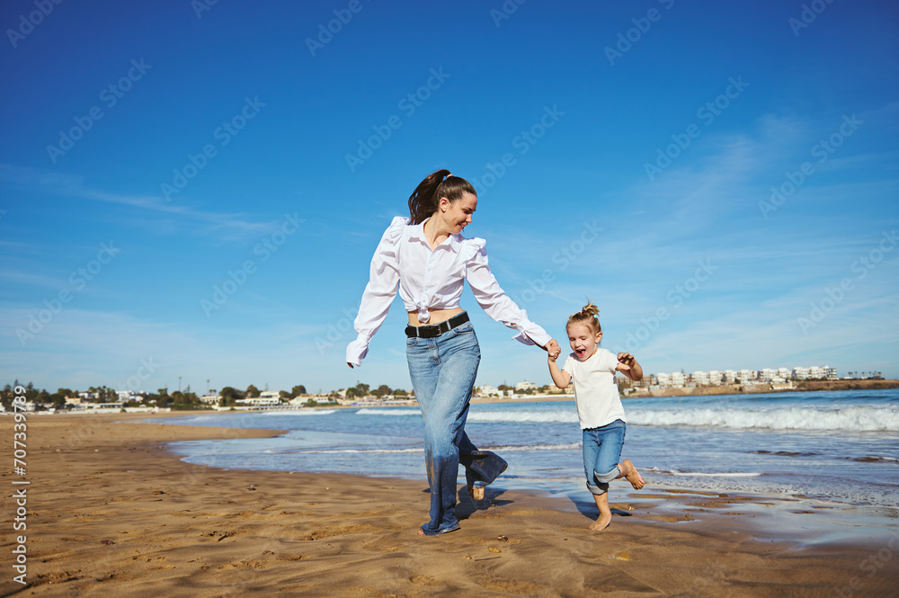 Young mother and her daughter running barefoot on warm water on waves, playing together, spending happy nice time Young mother and her daughter running barefoot on warm water on waves, playing together, spending happy nice time