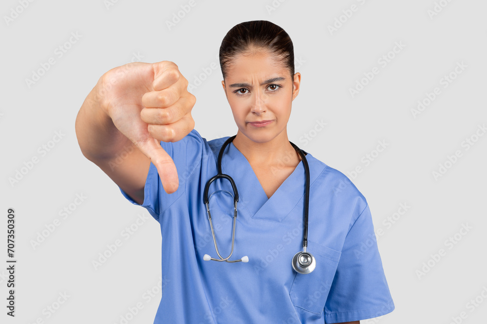A European millennial doctor in blue scrubs shows disapproval with a thumbs-down A European millennial doctor in blue scrubs shows disapproval with a thumbs-down