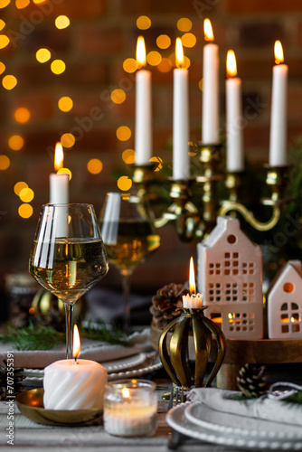 Luxury elegant Christmas dinner table setting with old vintage golden chandelier  candles  fir tree branches  wooden furniture. Porcelain houses as centrepiece  garland with lights on background