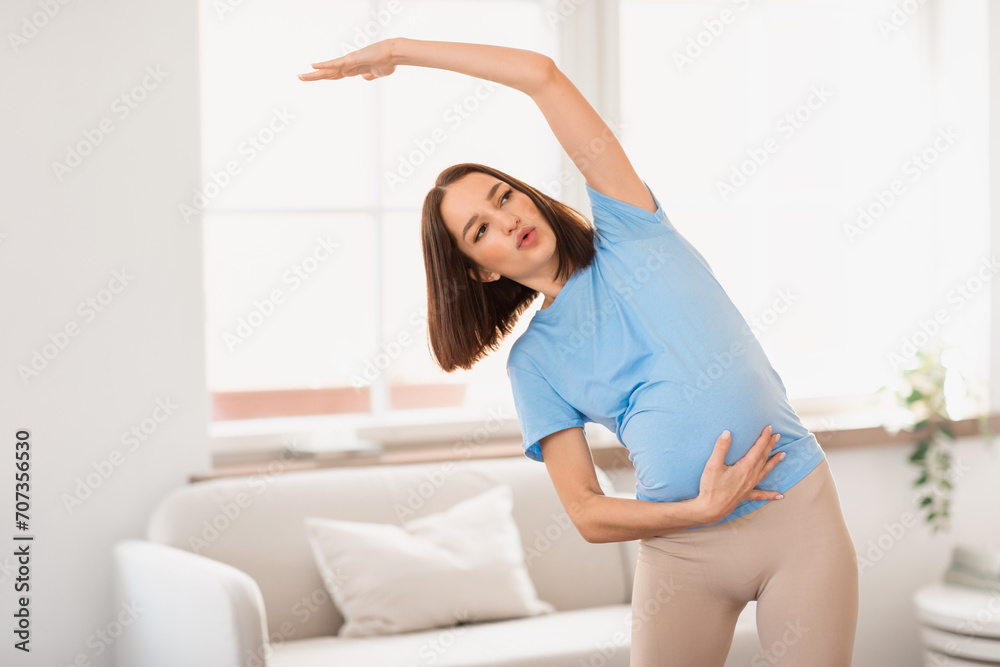 Millennial pregnant woman doing side bends with raised arm indoors Millennial pregnant woman doing side bends with raised arm indoors