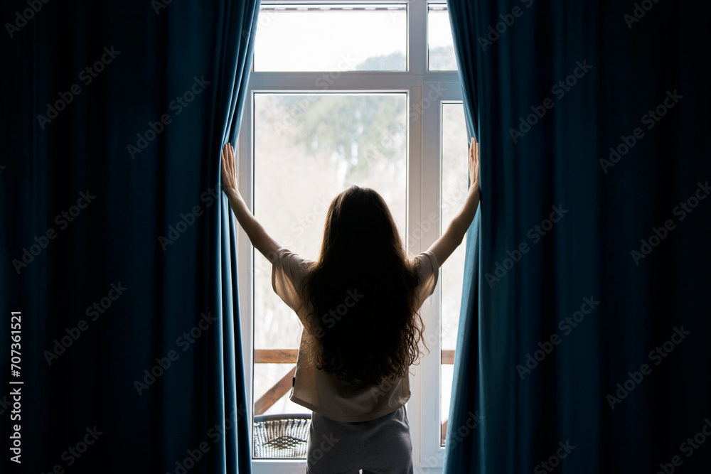 A woman with long hair stands between open curtains, welcoming natural light into a serene room through a large window, suggesting a sense of renewal or beginning. A woman with long hair stands between open curtains, welcoming natural light into a serene room through a large window, suggesting a sense of renewal or beginning.