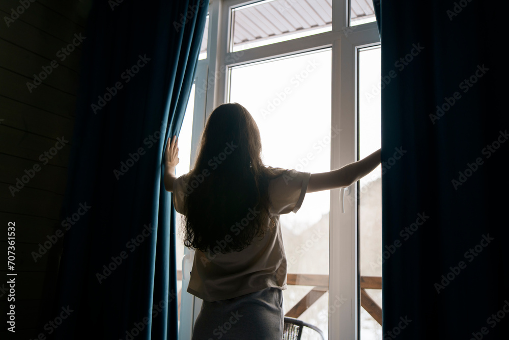 A woman with long hair stands between open curtains, welcoming natural light into a serene room through a large window, suggesting a sense of renewal or beginning.
