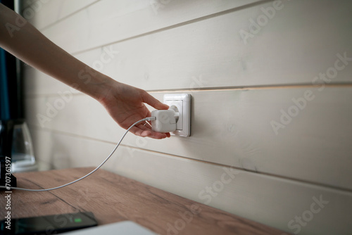 A persons hand is captured in the moment of inserting a two-pronged white electrical plug into a standard wall-mounted power socket, suggesting an indoor domestic or office setting. photo