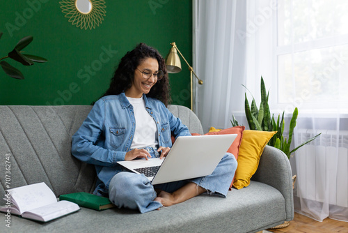 Young beautiful businesswoman freelancer working from home remotely, successful joyful Latin American woman sitting on sofa in living room at home, using laptop at work, typing on keyboard.