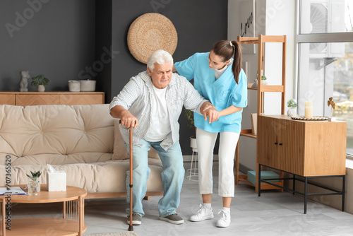 Nurse helping senior man with stick to stand up from sofa at home photo