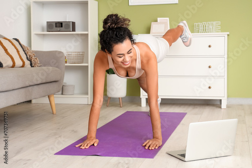 Young African-American woman training on mat with laptop in living room