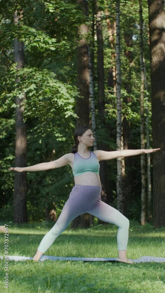 Vertical shot pregnant woman doing yoga standing in warrior position outdoors on sunny day. Young mother to be practice stretching yoga in green park with trees on background. Slide camera movement Vertical shot pregnant woman doing yoga standing in warrior position outdoors on sunny day. Young mother to be practice stretching yoga in green park with trees on background. Slide camera movement