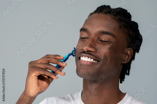 Cheerful handsome black man shaving his face with blue razor