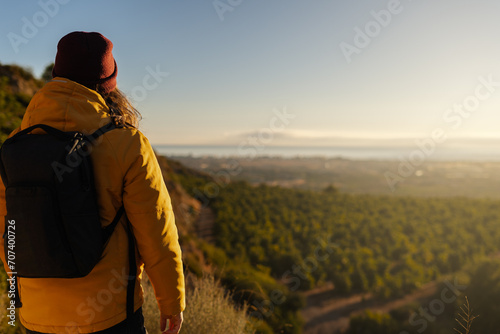 Rear view of a reflective hiker in a yellow jacket and red beanie standing on a trail and observing a scenic valley during golden hour.