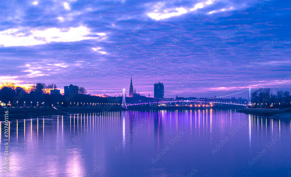 Cityscape and sunset over a river, with city lights and lit bridge and water reflections