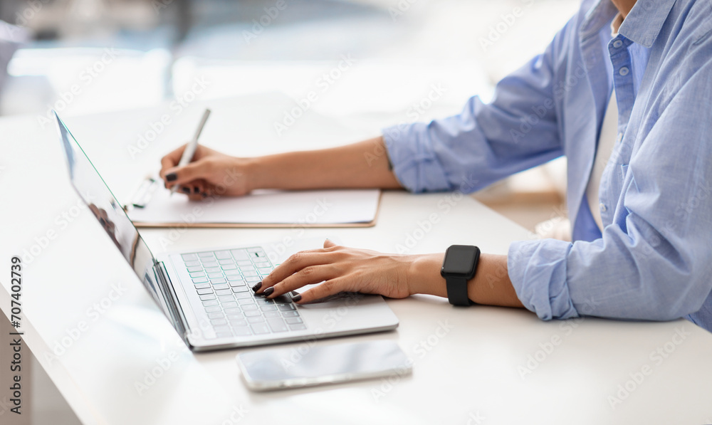 Professional woman in blue shirt multitasking, typing on a laptop while writing notes Professional woman in blue shirt multitasking, typing on a laptop while writing notes