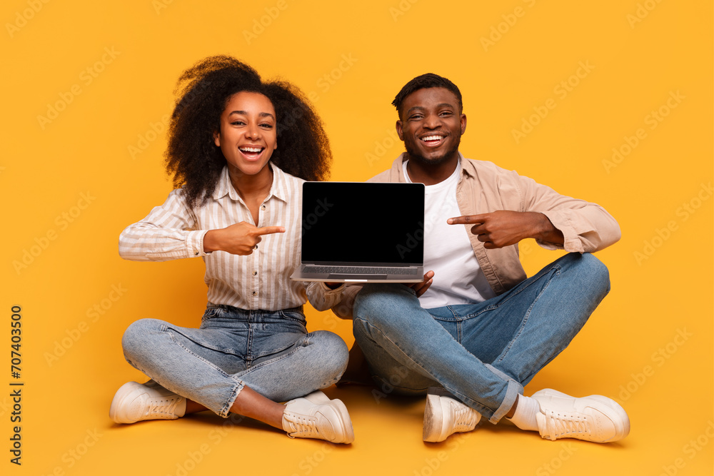Cheerful black couple pointing at blank laptop screen, seated on floor Cheerful black couple pointing at blank laptop screen, seated on floor