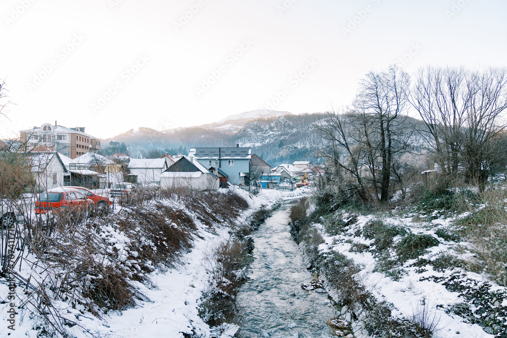 Mountain river flows between the snow-covered banks of a village at the foot of the mountains
