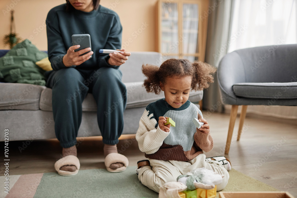 Full shot of African American girl with two ponytails playing with toys on living room floor while unrecognizable mother on sofa holding smartphone and thermometer - obrazy, fototapety, plakaty 