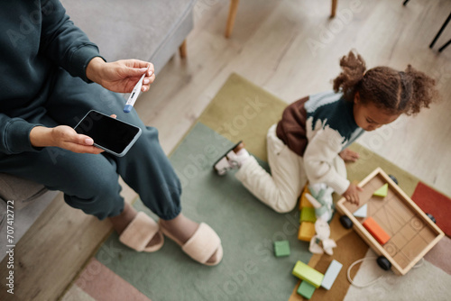Top down shot with focus on smartphone and digital thermometer in hands of unrecognizable mother while defocused girl child playing with toys on floor