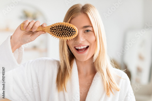 blonde young lady covers eye with wooden hair brush indoor